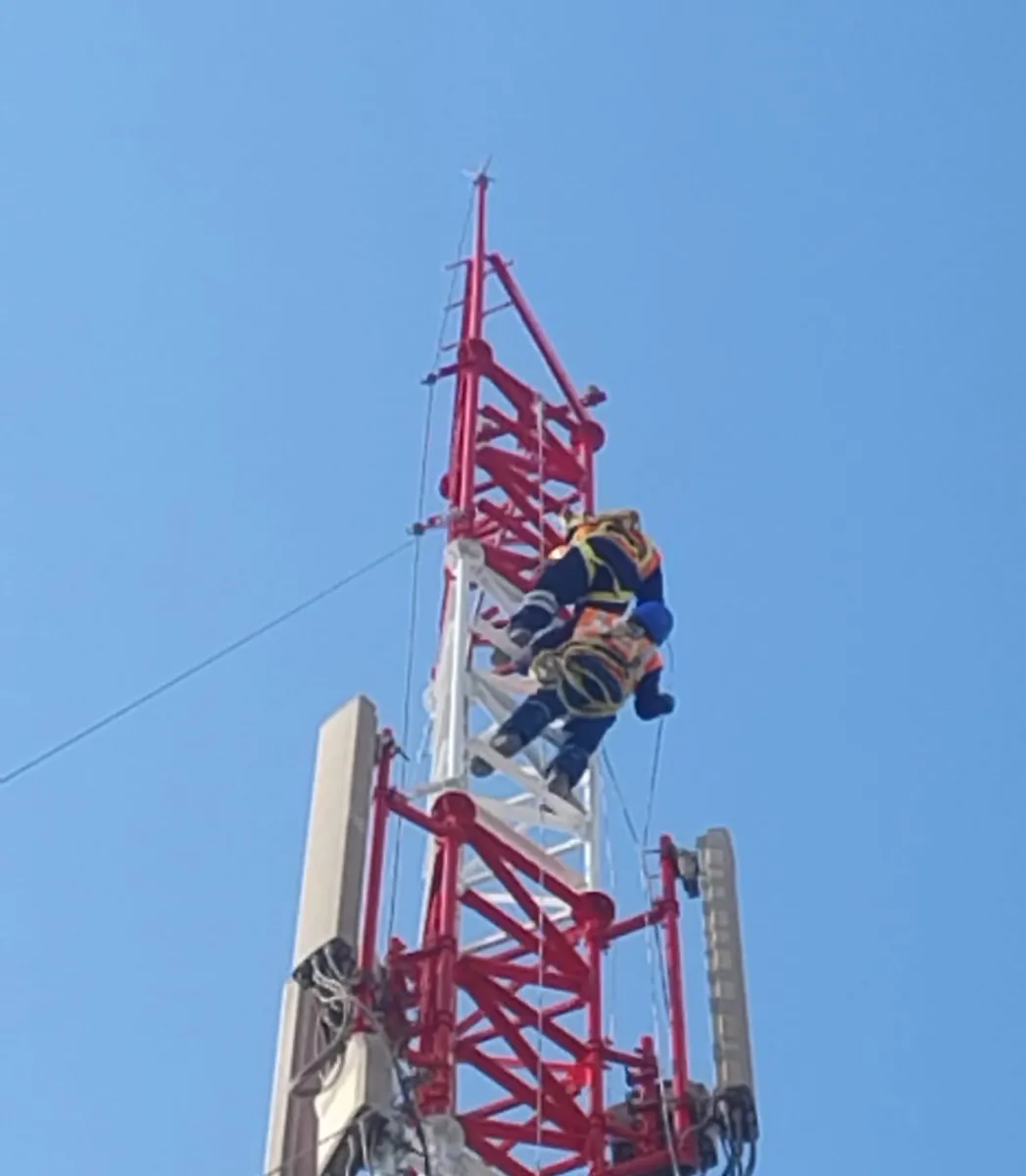 Técnico con arnés trabajando en lo alto de una torre roja y blanca con antenas.