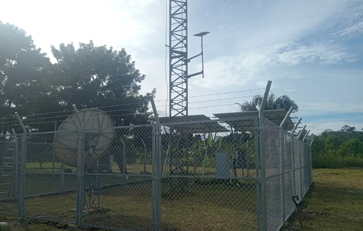 Cerco metálico de malla alrededor de una estación con torre, paneles solares y antena parabólica.
