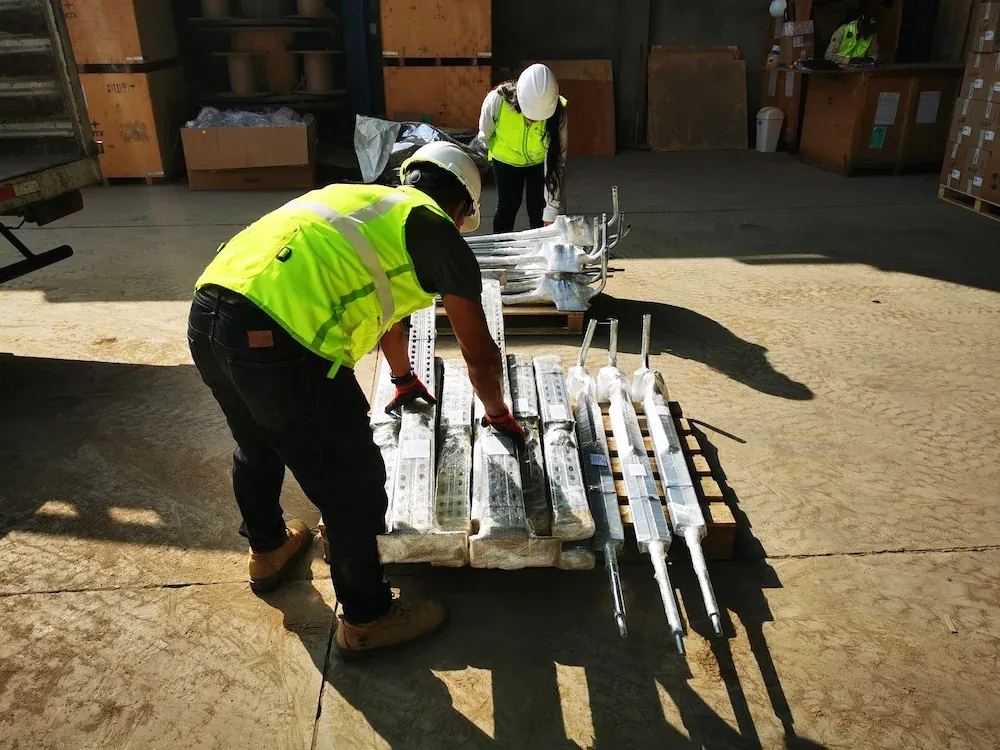 Dos trabajadores con casco y chaleco preparan en el almacén un pallet con piezas galvanizadas envueltas para su despacho.