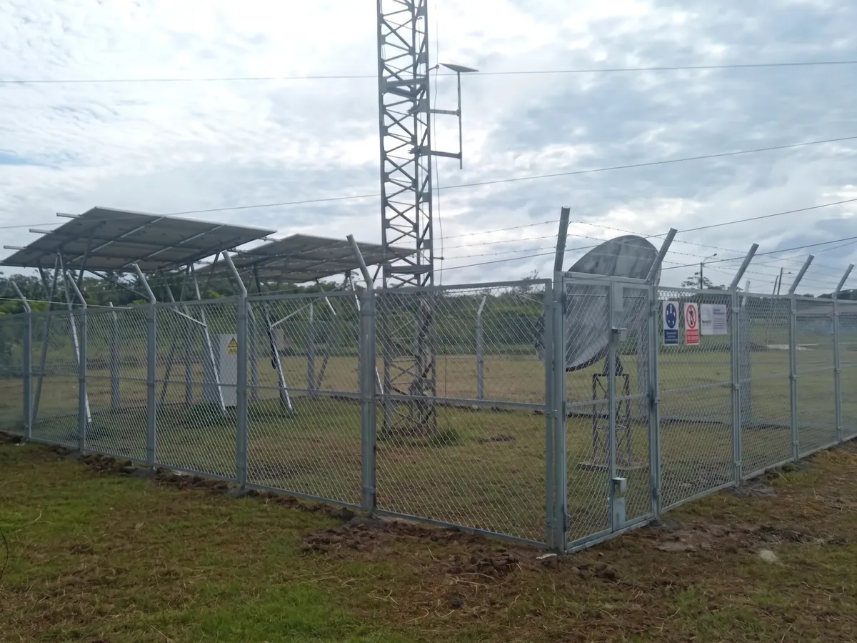 Estación de telecomunicaciones con su cerco de malla, paneles solares, antena parabólica y torre, sobre terreno con césped.