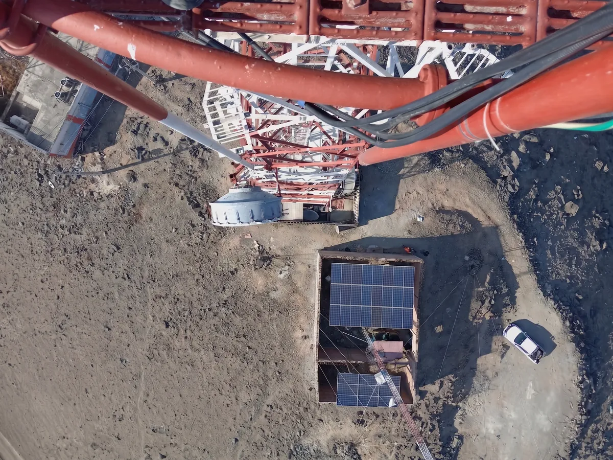 Vista desde lo alto de una torre hacia su base: la losa de la caseta con paneles solares y el terreno del sitio alrededor.