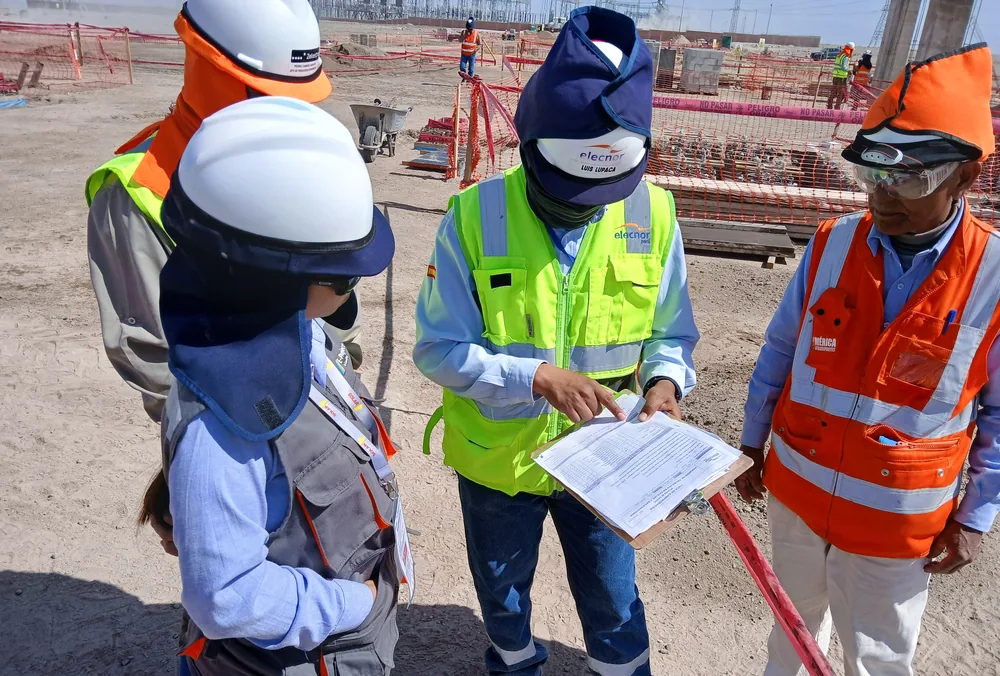 Cuatro personas con casco y chaleco revisando un documento en obra, con encofrados y estructuras al fondo.